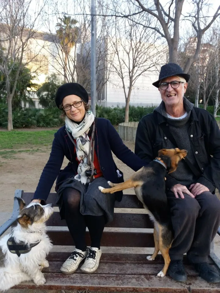 Fotografía de Karin du Croo y Miguel Gallardo sentados en un banco de parque con dos perros, ambos sonrientes y vestidos de manera informal con gorros negros.