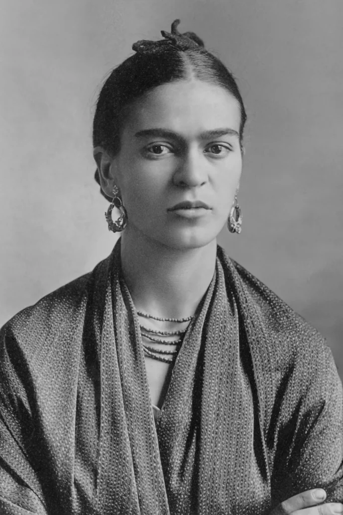 Retrato en blanco y negro de Frida Khalo, una mujer joven con el cabello recogido, mirada seria, pendientes grandes y un chal sobre los hombros, posando frente a un fondo neutro.