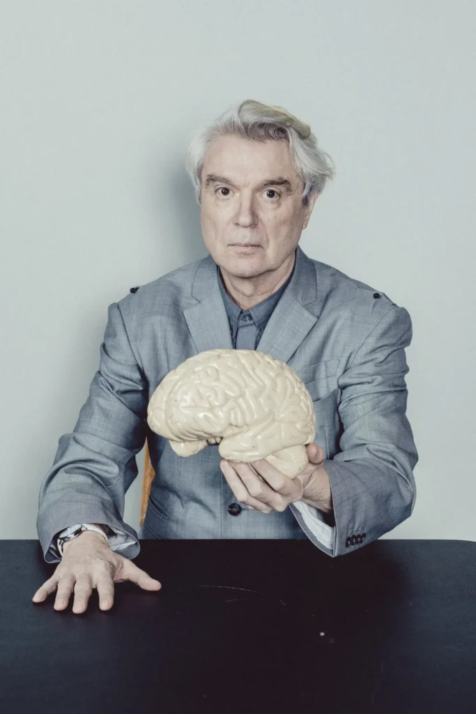 Retrato de David Byrne, un hombre de cabello canoso, vestido con traje gris, sentado frente a una mesa y sosteniendo un modelo anatómico de cerebro con expresión seria.