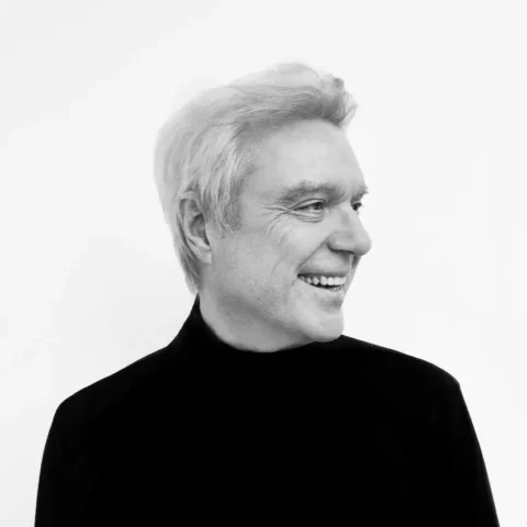 David Byrne en blanco y negro, cabello claro con jersey oscuro, sonriendo mientras mira hacia la derecha, sobre un fondo blanco.