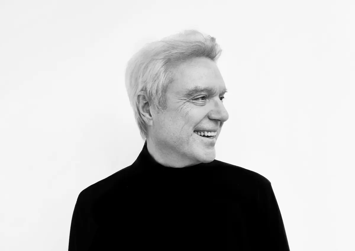David Byrne en blanco y negro, cabello claro con jersey oscuro, sonriendo mientras mira hacia la derecha, sobre un fondo blanco.