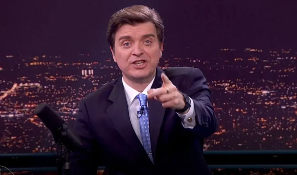 Marc Giró, presentador de televisión con traje oscuro, camisa blanca y corbata azul con lunares, señalando hacia la cámara durante un programa nocturno con una vista iluminada de ciudad al fondo.