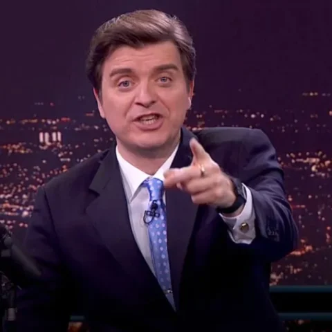 Marc Giró, presentador de televisión con traje oscuro, camisa blanca y corbata azul con lunares, señalando hacia la cámara durante un programa nocturno con una vista iluminada de ciudad al fondo.