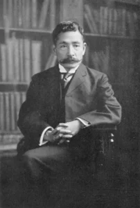 Fotografía en blanco y negro de Natsume Sōseki, escritor japonés, sentado con las manos entrelazadas, vistiendo traje y corbata, con una estantería de libros al fondo.