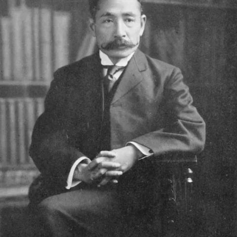 Fotografía en blanco y negro de Natsume Sōseki, escritor japonés, sentado con las manos entrelazadas, vistiendo traje y corbata, con una estantería de libros al fondo.