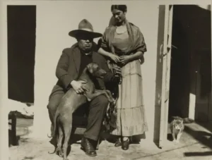 Fotografía antigua en blanco y negro de una pareja frente a una casa. El hombre está sentado con un perro sobre las piernas y lleva sombrero; la mujer está de pie a su lado, mirándolo, mientras un gato se asoma desde el interior de la casa.
