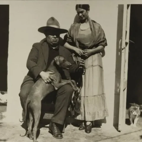 Fotografía antigua en blanco y negro de una pareja frente a una casa. El hombre está sentado con un perro sobre las piernas y lleva sombrero; la mujer está de pie a su lado, mirándolo, mientras un gato se asoma desde el interior de la casa.