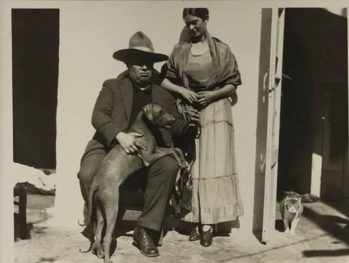 Fotografía antigua en blanco y negro de una pareja frente a una casa. El hombre está sentado con un perro sobre las piernas y lleva sombrero; la mujer está de pie a su lado, mirándolo, mientras un gato se asoma desde el interior de la casa.