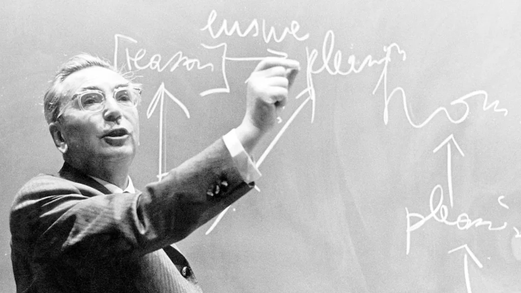 Fotografía en blanco y negro de Viktor Frankl escribiendo en una pizarra con tiza mientras gesticula durante una clase, con palabras y flechas visibles detrás de él.