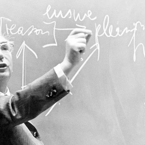 Fotografía en blanco y negro de Viktor Frankl escribiendo en una pizarra con tiza mientras gesticula durante una clase, con palabras y flechas visibles detrás de él.