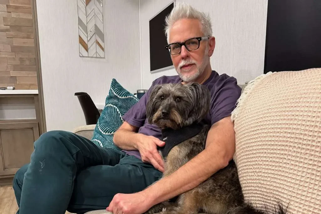 Hombre de cabello canoso y gafas negras sentado en un sofá moderno mientras sostiene un perro de pelaje oscuro en su regazo.