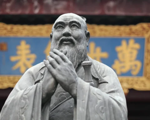 Escultura de bronce de un sabio con barba y túnica tradicional, con las manos unidas en gesto de respeto, frente a un fondo con caracteres chinos dorados sobre un cartel azul.