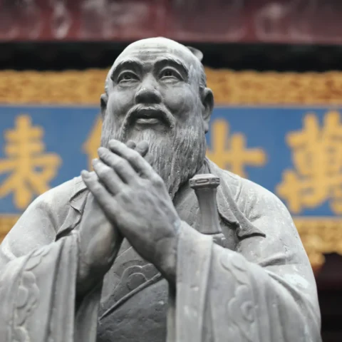 Escultura de bronce de un sabio con barba y túnica tradicional, con las manos unidas en gesto de respeto, frente a un fondo con caracteres chinos dorados sobre un cartel azul.