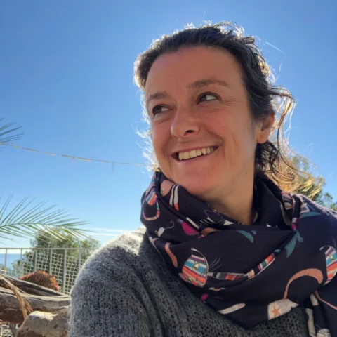 Retrato María Casaoliva sonriendo al sol en un día despejado, con bufanda ilustrada y jersey gris, con el cielo azul de fondo.