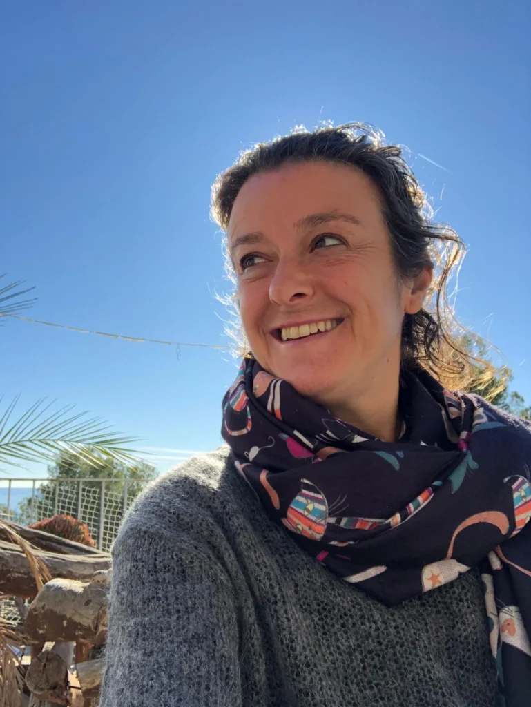 Retrato María Casaoliva sonriendo al sol en un día despejado, con bufanda ilustrada y jersey gris, con el cielo azul de fondo.