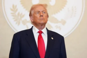 Donald Trump, presidente de EEUU vestido con traje oscuro y corbata roja, posando con gesto serio frente a un fondo blanco con un emblema circular