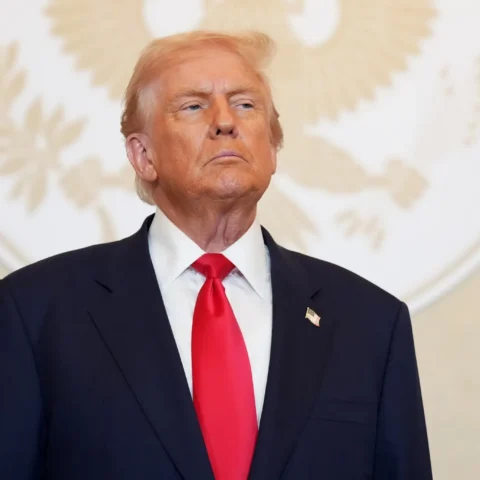 Donald Trump, presidente de EEUU vestido con traje oscuro y corbata roja, posando con gesto serio frente a un fondo blanco con un emblema circular