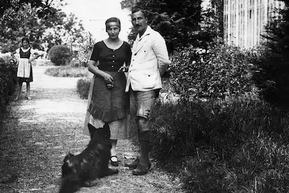 Fotografía antigua en blanco y negro de una pareja posando en un sendero ajardinado mientras un perro pequeño corre frente a ellos; al fondo, una niña observa la escena.