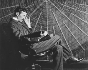 Nikola Tesla, sentado en una silla de despacho, acariciando un gato blanco y negro frente a una de sus invenciones eléctricas