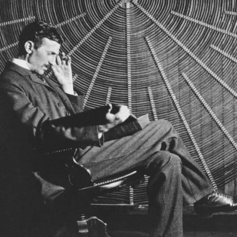 Nikola Tesla, sentado en una silla de despacho, acariciando un gato blanco y negro frente a una de sus invenciones eléctricas