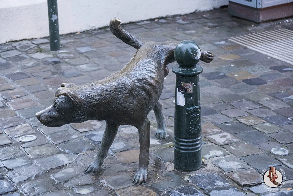 Escultura de bronce de un perro levantando la pata para orinar junto a un bolardo en una calle empedrada de Bruselas.