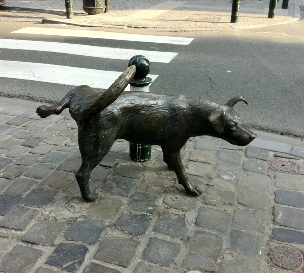 Escultura de bronce de un perro levantando la pata para orinar junto a un bolardo en una calle empedrada de Bruselas.
