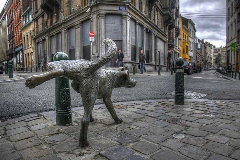 Escultura de bronce de un perro levantando la pata para orinar junto a un bolardo en una calle empedrada de Bruselas.