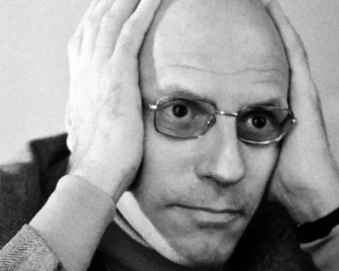 Retrato en blanco y negro del filósofo Michel Foucault con gafas, sosteniendo la cabeza con ambas manos y mirando fijamente a la cámara.
