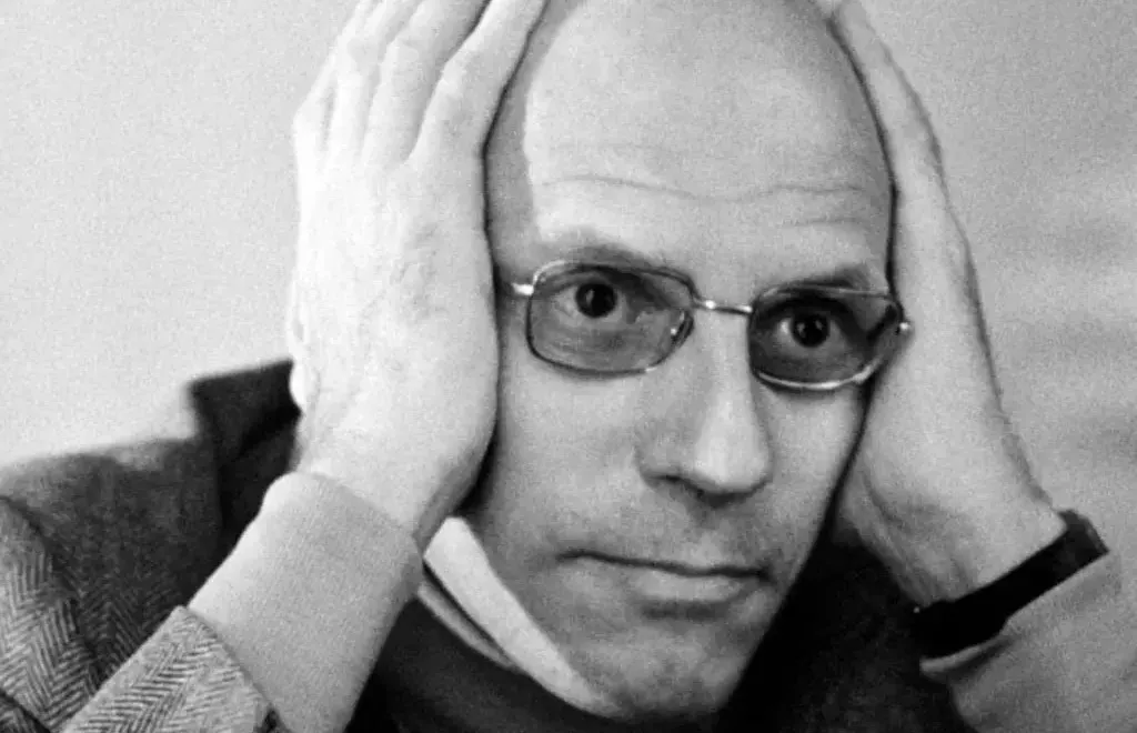 Retrato en blanco y negro del filósofo Michel Foucault con gafas, sosteniendo la cabeza con ambas manos y mirando fijamente a la cámara.