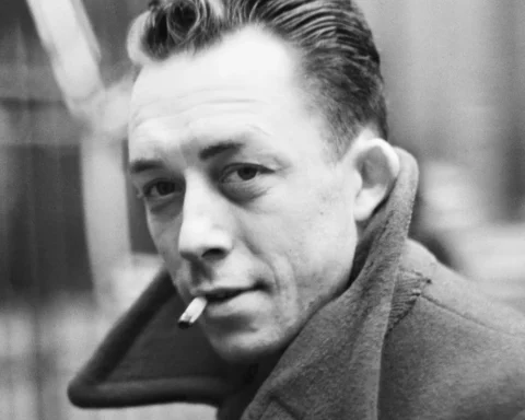 Retrato en blanco y negro de un Albert Camus con abrigo oscuro y mirada seria, apoyado en una pared, en un ambiente urbano sobrio.