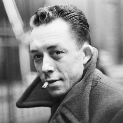 Retrato en blanco y negro de un Albert Camus con abrigo oscuro y mirada seria, apoyado en una pared, en un ambiente urbano sobrio.