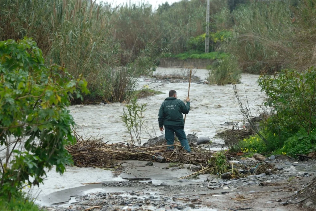 Agente de la Guardia Civil camina con una vara por el cauce de un río crecido, rodeado de vegetación y restos arrastrados por la corriente tras una crecida.