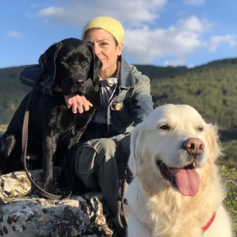 Sara Escudero sentada en un prado abrazando a un perro golden retriever entre flores silvestres.