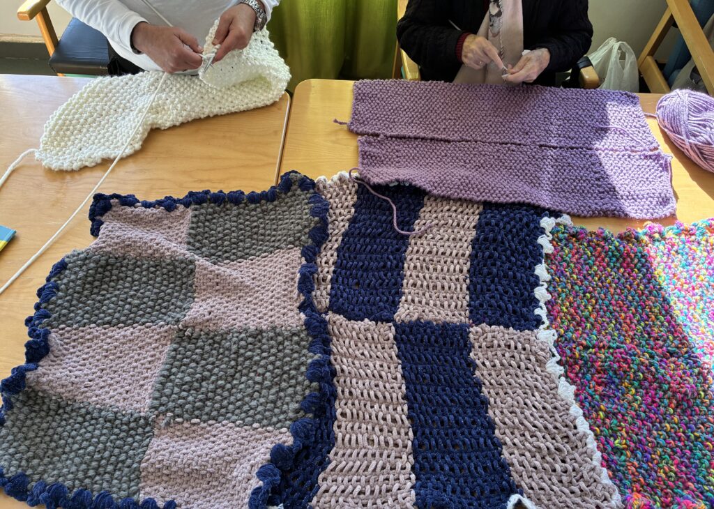 Personas tejiendo mantas de lana de colores sobre una mesa, con varias piezas hechas a mano en primer plano.