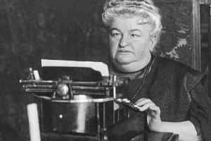 Retrato en blanco y negro de Emilia Pardo Bazán sentada ante una máquina de escribir, concentrada mientras sostiene el papel con una mano