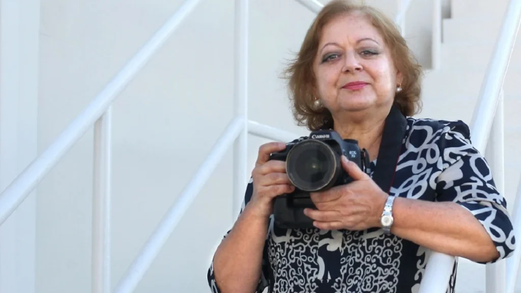 Retrato en color de Cristina García Rodero con su cámara Canon, apoyada en una escalera blanca.