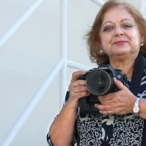 Retrato en color de Cristina García Rodero con su cámara Canon, apoyada en una escalera blanca.