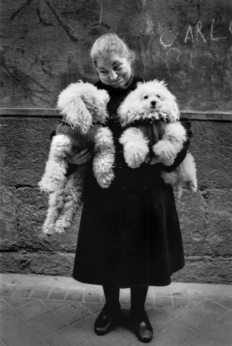 Retrato en blanco y negro de una mujer sonriente sosteniendo en brazos a dos perros caniche blancos.