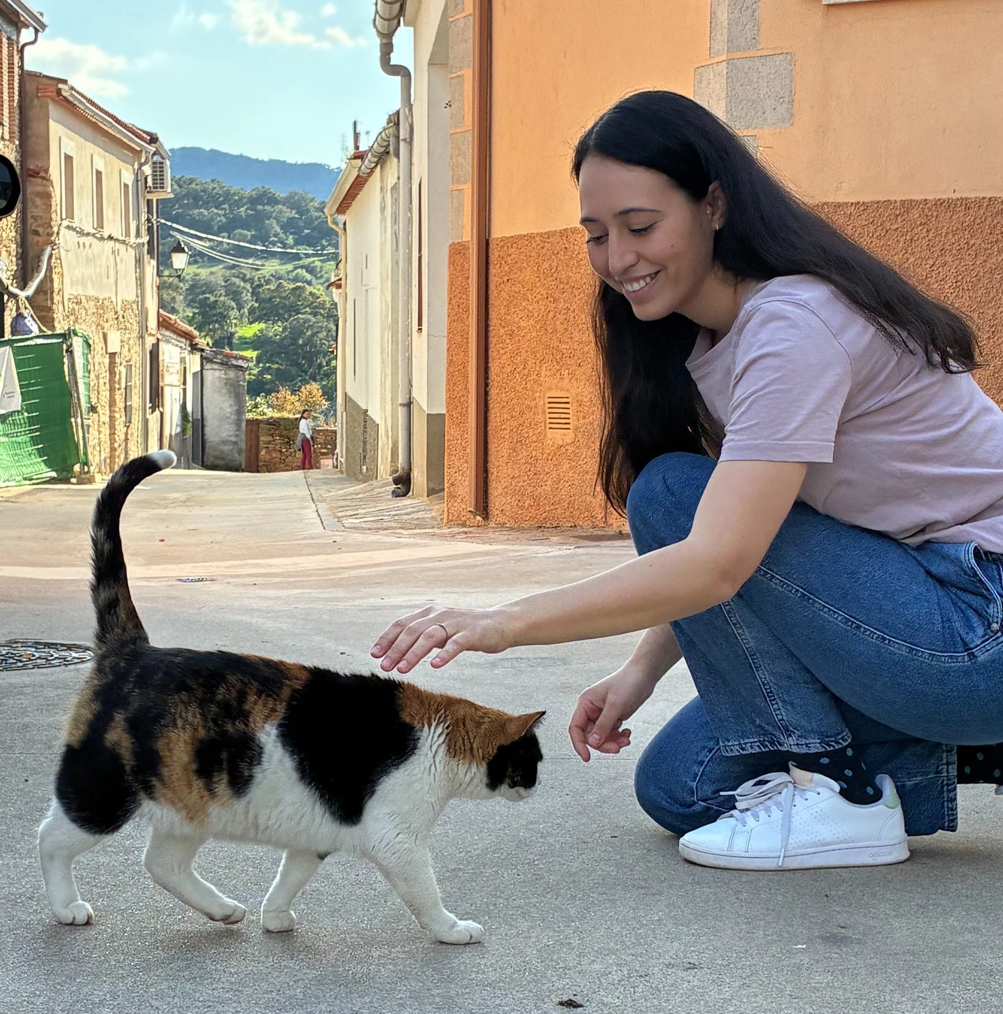 Sara Ramiro acariciando a un gato tricolor en una calle de pueblo con casas tradicionales al fondo.