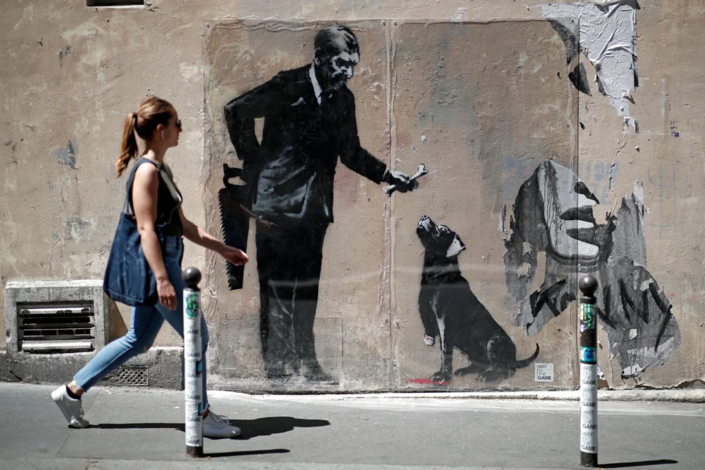 Intervención de arte urbano en una pared que muestra una escena figurativa con un personaje y un perro, utilizando tonos oscuros y estilo stencil característico del arte callejero.