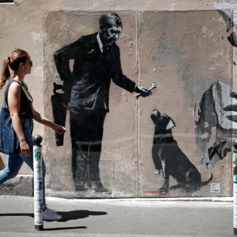 Intervención de arte urbano en una pared que muestra una escena figurativa con un personaje y un perro, utilizando tonos oscuros y estilo stencil característico del arte callejero.