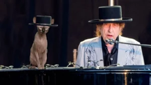 Escena de Bob Dylantocando el piano con sombrero negro mientras canta al micrófono; a su lado, un gato esfinge también lleva un sombrero similar sobre el piano.