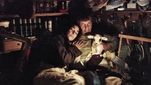 Escena cinematográfica en un interior oscuro donde una pareja sostiene un cordero iluminado por una pequeña llama, rodeados de objetos en estanterías.