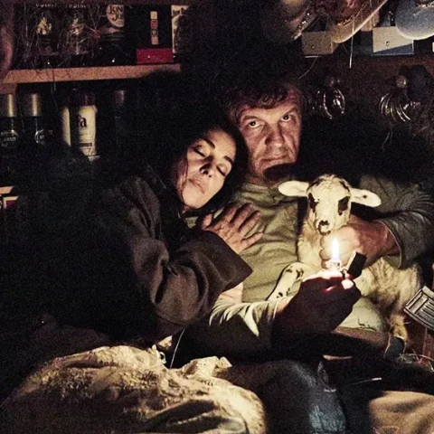 Escena cinematográfica en un interior oscuro donde una pareja sostiene un cordero iluminado por una pequeña llama, rodeados de objetos en estanterías.