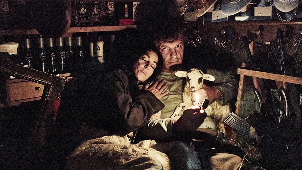 Escena cinematográfica en un interior oscuro donde una pareja sostiene un cordero iluminado por una pequeña llama, rodeados de objetos en estanterías.