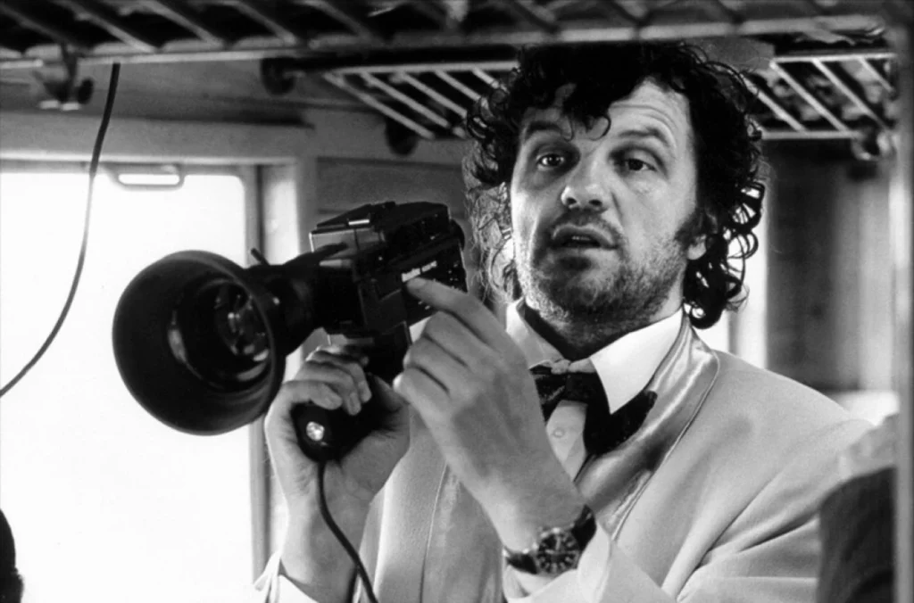 Fotografía en blanco y negro de Emir Kusturica sosteniendo una cámara de cine, vestido con traje y pajarita, en un entorno interior.