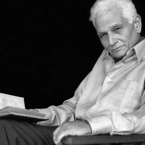 Retrato en blanco y negro de Jacques Derrida sentado en una silla, con un libro abierto en la mano y una vela encendida sobre una mesa a su lado, en un ambiente íntimo y reflexivo.