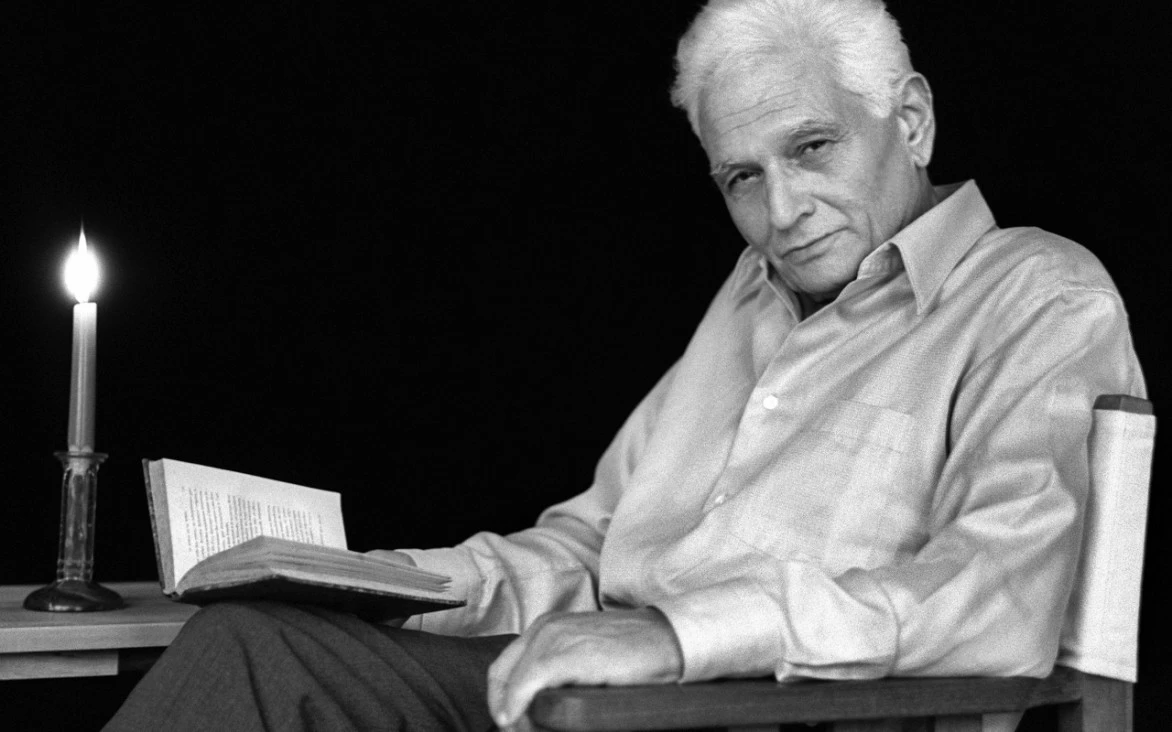 Retrato en blanco y negro de Jacques Derrida sentado en una silla, con un libro abierto en la mano y una vela encendida sobre una mesa a su lado, en un ambiente íntimo y reflexivo.