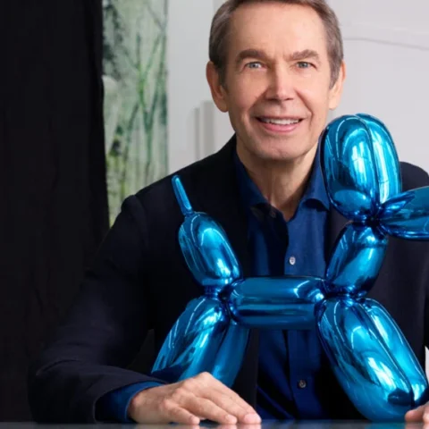 Jeff Koons posando junto a una escultura azul con forma de perro globo en un estudio de arte.