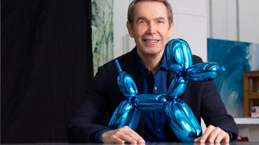 Jeff Koons posando junto a una escultura azul con forma de perro globo en un estudio de arte.